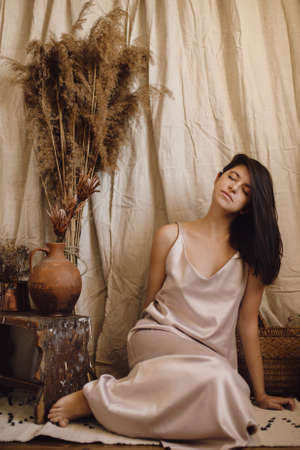 Stylish fashionable woman in silk dress sensually posing in boho style room with dry grass and flowers on beige background. Modern bohemian bride. Young attractive female in atmospheric interiorの写真素材