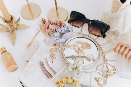 Modern summer accessories. Golden jewellery, sunglasses, hair clips, cosmetics, perfume and lace lingerie on white table with vintage candles and boho mirror. Feminine essentialsの写真素材