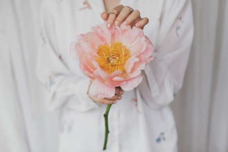 Sensual beautiful woman with pink peony in hands. Young stylish female in boho floral shirt gently holding big peony flower. Tender soft image. Spring aestheticsの写真素材