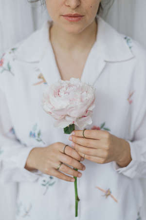 Beautiful stylish woman holding pink peony. Young female in boho floral shirt gently holding big peony flower in hands. Sensual soft image. Spring aestheticsの写真素材