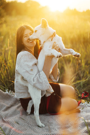 Stylish happy woman playing with her white dog on blanket in warm sunny light in summer meadow. Summer vacation and picnic with pet. Young boho female having fun with swiss shepherd puppyの写真素材