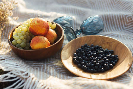 Fresh delicious fruits in wooden plates and blue wineglasses on yellow plaid in warm sunny light in summer meadow. Summer picnic concept. Vacation and leisureの写真素材