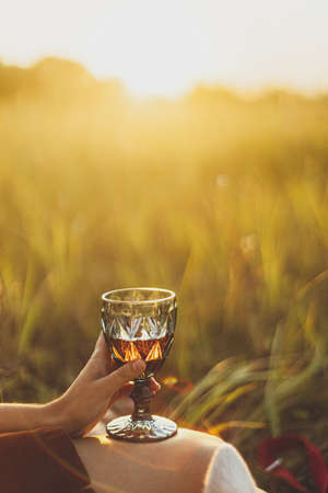 Wine glass in woman hand close up on background of summer meadow grass in warm sunset light.  Summer vacation and picnic. Stylish woman relaxing with glass of wine and white dog.の写真素材