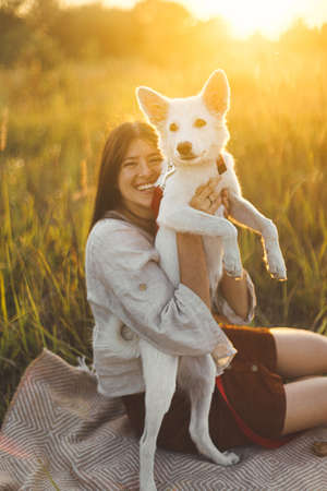 Stylish happy woman playing with her white dog on blanket in warm sunny light in summer meadow. Summer vacation and picnic with pet. Young boho female having fun with swiss shepherd puppyの写真素材