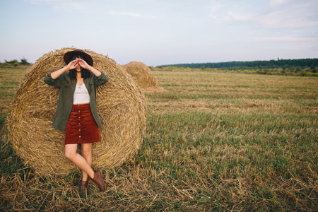 Stylish woman in hat standing at hay bale in summer evening in field. Atmospheric tranquil moment. Young fashionable female relaxing at haystack, summer vacation in countrysideの写真素材