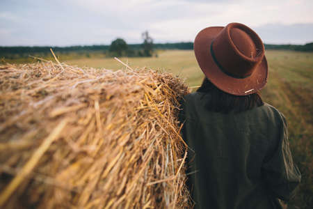 Stylish woman in hat with straw standing at haystack in summer evening field, back view. Atmospheric tranquil moment in countryside. Young female resting at hay bale.の写真素材