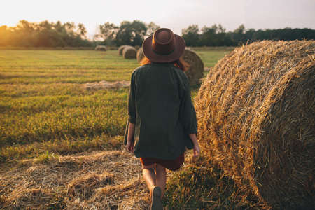 Beautiful stylish woman in hat walking at haystacks in sunset light in summer field. Atmospheric tranquil moment in countryside. Young female enjoying evening at hay bale in warm sunshineの写真素材