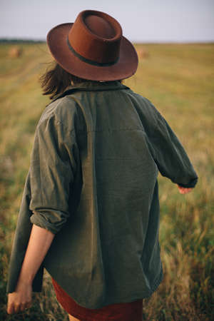 Beautiful carefree woman in hat walking in evening summer field. Young happy stylish female relaxing in countryside, dancing and enjoying evening. Atmospheric moment. Back viewの写真素材