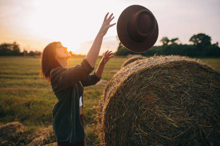 Beautiful carefree woman catching hat in sunset light at haystacks in evening summer field. Young...