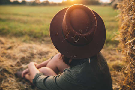 Stylish woman in hat looking at sunset light, sitting at haystacks in summer field. Atmospheric tranquil moment in countryside. Young female enjoying evening at hay bale in warm sunshineの写真素材
