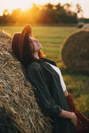 Beautiful stylish woman in hat relaxing on haystacks in sunset light in summer field. Atmospheric tranquil moment in countryside. Young female enjoying evening at hay bale in warm sunshineの写真素材