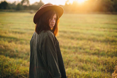 Beautiful stylish woman in hat posing at haystacks in sunset light in summer field. Atmospheric tranquil moment in countryside. Young female relaxing at hay bale in evening warm sunshineの写真素材