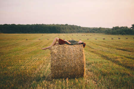 Beautiful carefree woman lying on haystack enjoying evening in summer field. Young happy female relaxing on hay bale in countryside. Atmospheric tranquil momentの写真素材