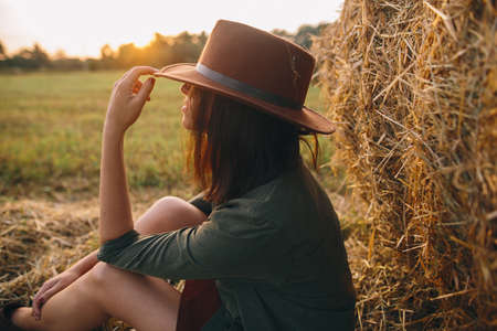 Beautiful stylish woman in hat enjoying sunset, sitting at haystacks in evening summer field. Atmospheric tranquil moment in countryside. Young fashionable female relaxing at hay bale in sunshineの写真素材