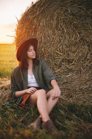 Beautiful stylish woman in hat sitting at haystacks enjoying evening in sunny summer field. Atmospheric tranquil moment in countryside. Young female relaxing at hay bale in warm sunsetの写真素材