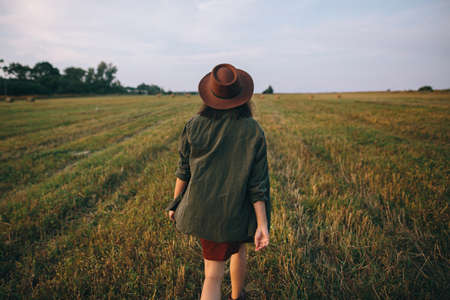 Beautiful carefree woman in hat walking in evening summer field. Young happy stylish female relaxing in countryside, dancing and enjoying evening. Atmospheric moment. Back viewの写真素材