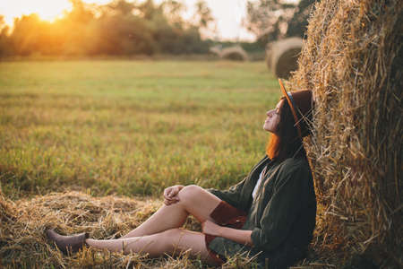 Beautiful stylish woman in hat enjoying sunset, sitting at haystacks in evening summer field. Atmospheric tranquil moment in countryside. Young fashionable female relaxing at hay bale in sunshineの写真素材