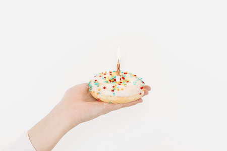 Happy Birthday. Hand holding delicious birthday donut with candle on background of white wall in room. Birthday party celebration. Colorful doughnut with sprinkles and gold candleの写真素材