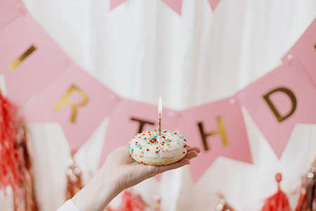 Happy Birthday. Hand holding delicious birthday donut with candle on background of pink garland and decorations in room.  Birthday party celebration. Colorful doughnut with sprinklesの写真素材