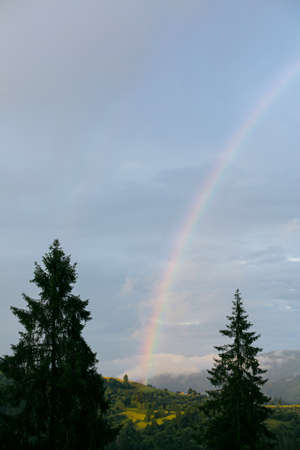 Rainbow in mountains after rain. Beautiful view on mountain hill with rainbow among trees from cloudy sunny sky in evening. Traveling and exploring Carpathians Mountainsの写真素材