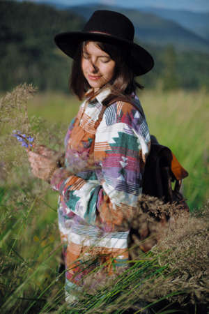 Happy woman traveler with backpack and hat gathering wildflowers on mountain hills and relaxing. Wanderlust and travel concept. Stylish female hiking in mountains, tranquil momentの写真素材