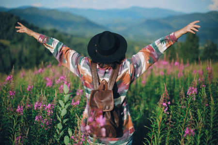 Woman traveler with backpack and hat standing in beautiful wildflowers and holding hands up on...