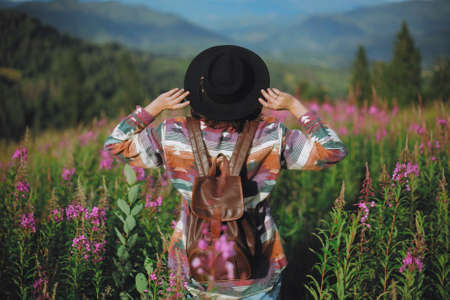 Wanderlust and travel concept. Woman traveler with backpack and hat standing in beautiful wildflowers and looking on mountain hills.  Stylish female hiking in mountains, atmospheric momentの写真素材