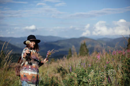 Happy woman traveler with backpack and hat gathering wildflowers on mountain hills and relaxing. Wanderlust and travel concept. Stylish female hiking in mountains, tranquil momentの写真素材