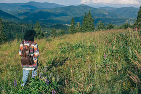 Woman traveler with backpack and hat standing on beautiful hill and looking on mountains. Wanderlust and travel concept. Stylish female hiking in mountains, atmospheric moment. Space for textの写真素材