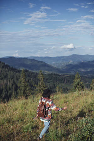 Woman traveler with backpack and hat walking on beautiful mountain hills and gathering wildflowers. Wanderlust and travel concept. Stylish female hiking in mountains, atmospheric momentの写真素材