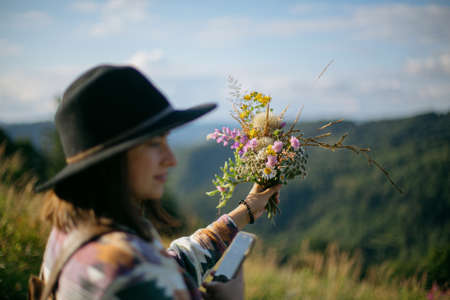 Woman traveler in hat holding bouquet of wildflowers on background of mountain hills and sky. Stylish female hiking in mountains, gathering flowers. Wanderlust and travel concept.の写真素材