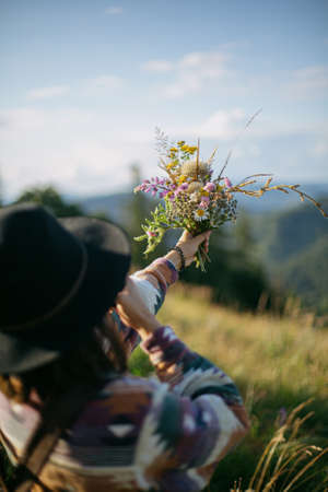 Woman traveler in hat holding bouquet of wildflowers on background of mountain hills and sky. Stylish female hiking in mountains, gathering flowers. Wanderlust and travel concept.の写真素材