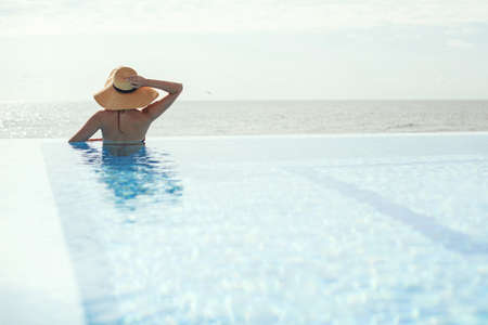 Summer vacation. Beautiful woman in hat relaxing at edge in blue pool, enjoying calm sea view. Slim...