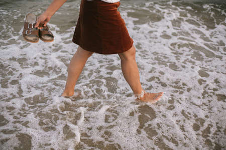 Carefree hipster woman walking barefoot in sea waves on beach with flip flops in hand. Feet in water...