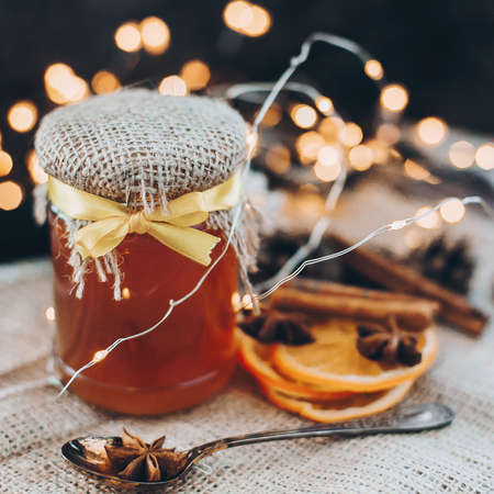 Honey in glass jar with ribbon and spoon with anise star, cinnamon, dried oranges on background of...