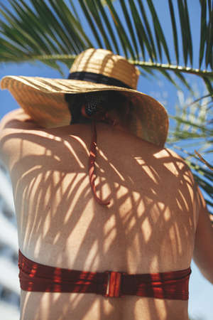 Shadow of palm leaves on back of beautiful woman in hat relaxing at pool enjoying summer vacation....