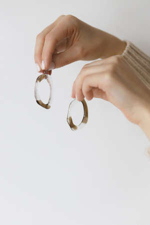 Hands holding stylish modern earrings on background of white wall. Unusual fashionable fused glass earrings in female hands with white manicure. Contemporary giftの写真素材