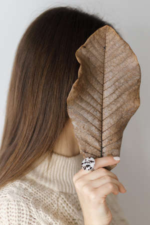 Beautiful stylish woman hiding face with big brown leaf with hand with modern round ring....