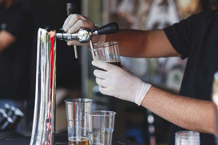 Barman pouring fresh beer in plastic cup at street food festival, close up. Man hands in gloves pouring refreshing drink at counter in bar or pub. Summer vacation picnic. Street food marketの写真素材