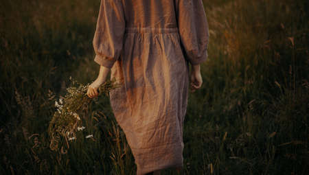 Beautiful woman in linen dress walking with wildflowers in hand in summer meadow in sunset. Atmospheric stylish vintage image. Young female in rustic dress relaxing in evening in countrysideの写真素材