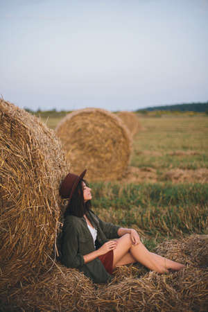 Beautiful stylish woman in hat sitting at hay bale in summer evening field. Happy young female relaxing at haystacks, summer vacation in countryside. Tranquility, countryside slow lifeの写真素材