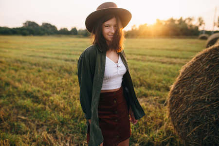 Beautiful stylish woman in hat walking at haystacks in sunset light in summer field. Atmospheric...