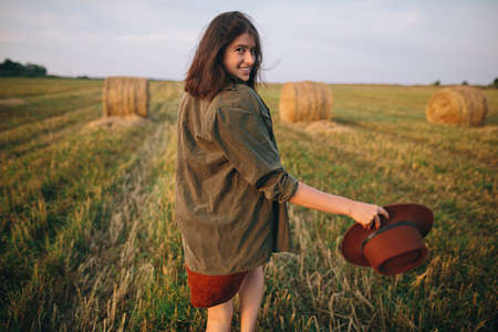 Beautiful carefree woman in hat walking and smiling in evening summer field. Young happy stylish female relaxing in countryside, enjoying evening. Atmospheric moment.の写真素材