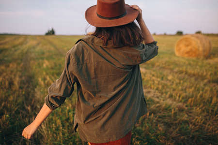 Beautiful carefree woman in hat walking in evening summer field. Young happy stylish female relaxing in countryside, dancing and enjoying evening. Atmospheric moment. Back viewの写真素材