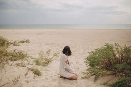 Beautiful woman with windy hair sitting on sandy beach on background of green grass and sea, calm...