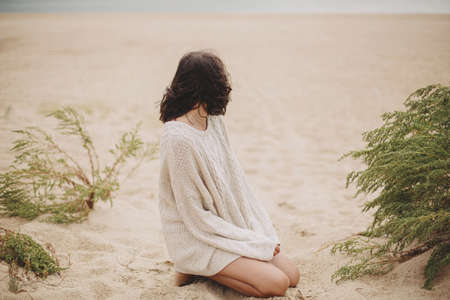 Beautiful woman sitting on sandy beach on background of green grass and looking at sea, calm tranquil moment. Stylish young female in beige knitted sweater relaxing on windy cold beachの写真素材