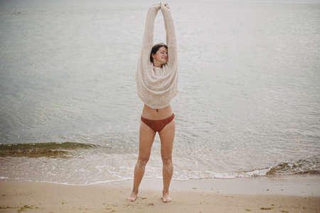 Carefree beautiful woman with windy hair in knitted sweater and bikini standing on sandy beach at...