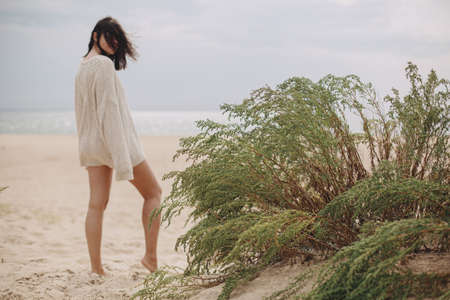 Green grass on sandy beach on background of blurred silhouette of beautiful woman with windy hair, calm tranquil moment. Stylish young female in beige knitted sweater relaxing on coastの写真素材
