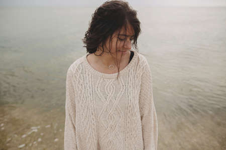 Carefree beautiful woman with curly hair posing on background of  beach and cold sea, tranquil moment. Portrait of stylish young female in knitted sweater relaxing and enjoying day on coastの写真素材