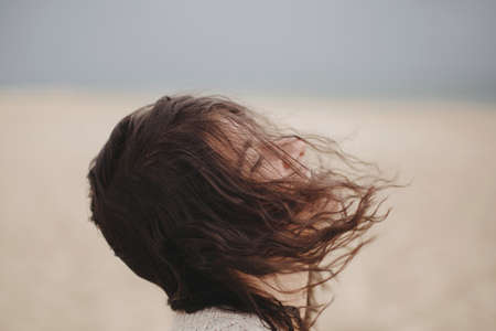 Carefree beautiful woman with windy hair on background of sandy beach and sea, tranquil moment. Portrait of stylish young female relaxing and enjoying vacation on coastの写真素材